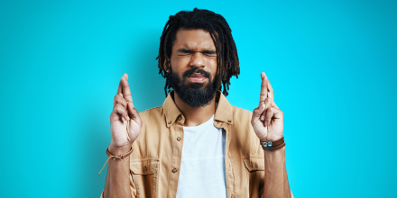 Young African man in casual clothing keeping fingers crossed while standing against blue background